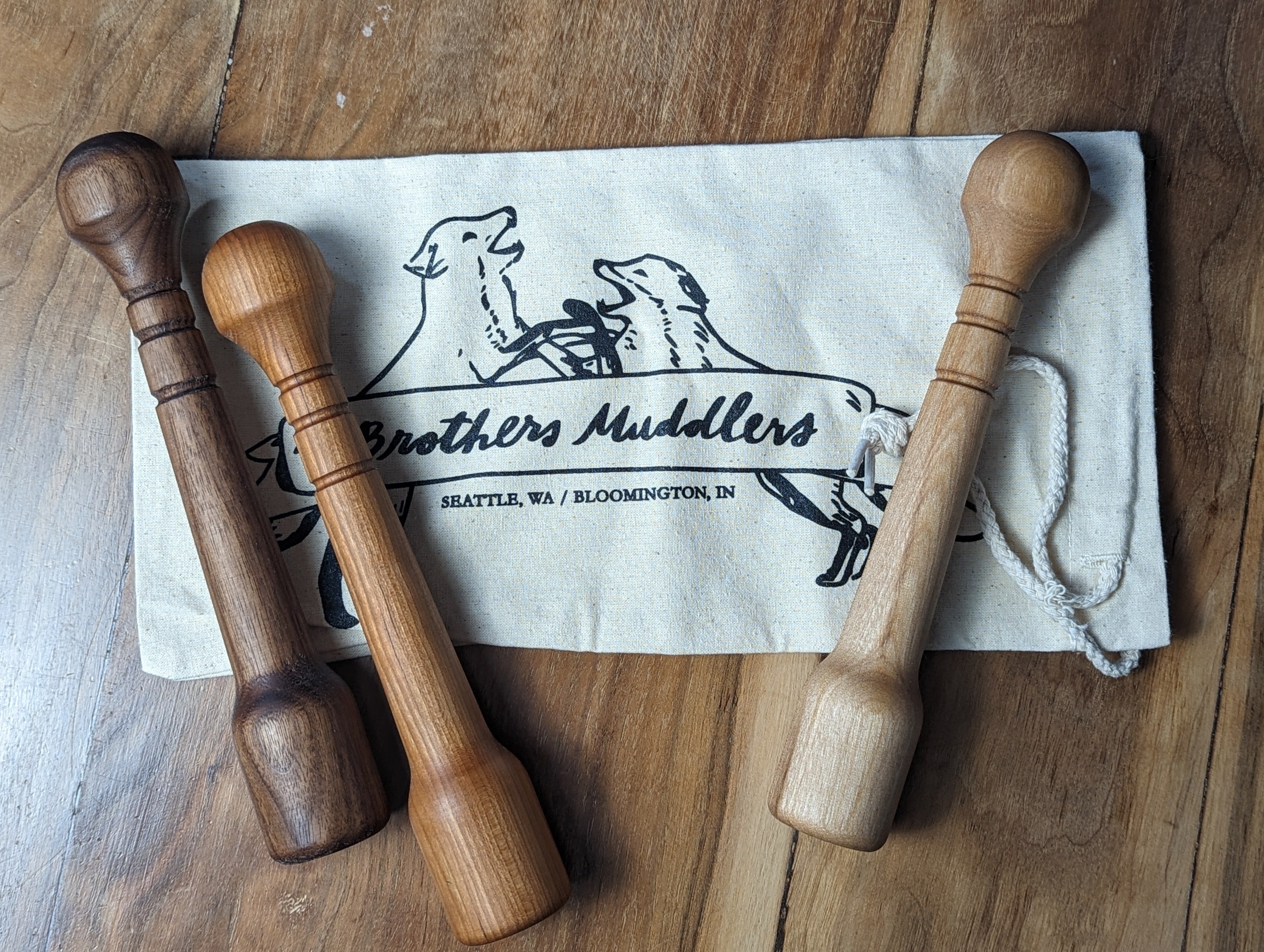 Three wooden muddlers on a cloth with 'Brothers Muddlers' branding on a wooden surface.